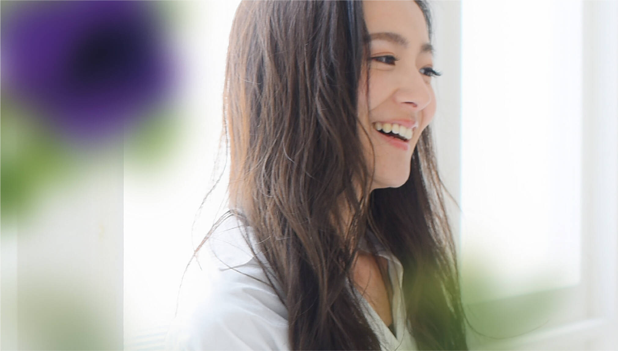 白いシャツを着た女性が笑顔で横を向く。柔らかい光が差し込む室内でリラックスした雰囲気。